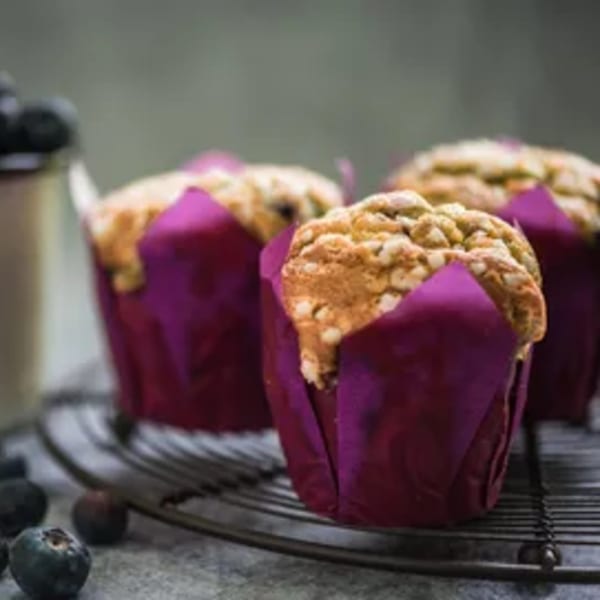 Three blueberry muffins in purple wrappers on a cooling rack, with fresh blueberries scattered nearby.