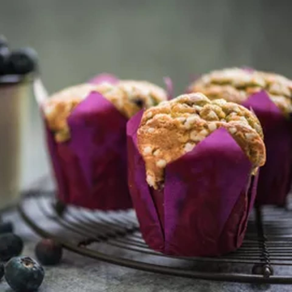 Three blueberry muffins in purple wrappers on a cooling rack, with fresh blueberries scattered nearby.