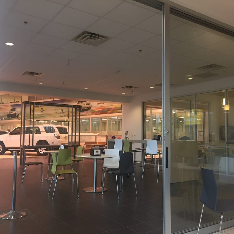 Empty dining area with tables and chairs, with a car dealership visible through a glass wall.