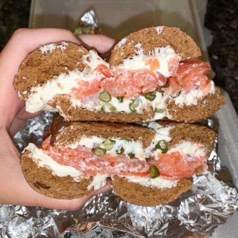 A New York Bagel Company bagel with lox, cream cheese, and capers, held over foil.