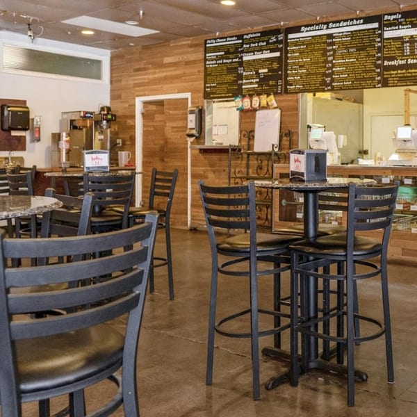 Interior of New York Bagel Company with high-top tables, chairs, and menu boards on a wood-paneled wall.